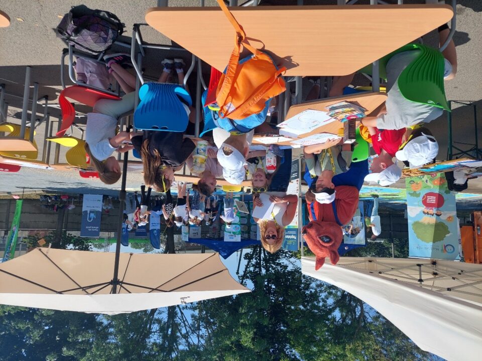 Mókusjelmezben egy ember, mellette egy másik ember és gyerekek ülnek asztaloknál egy rendezvényen.